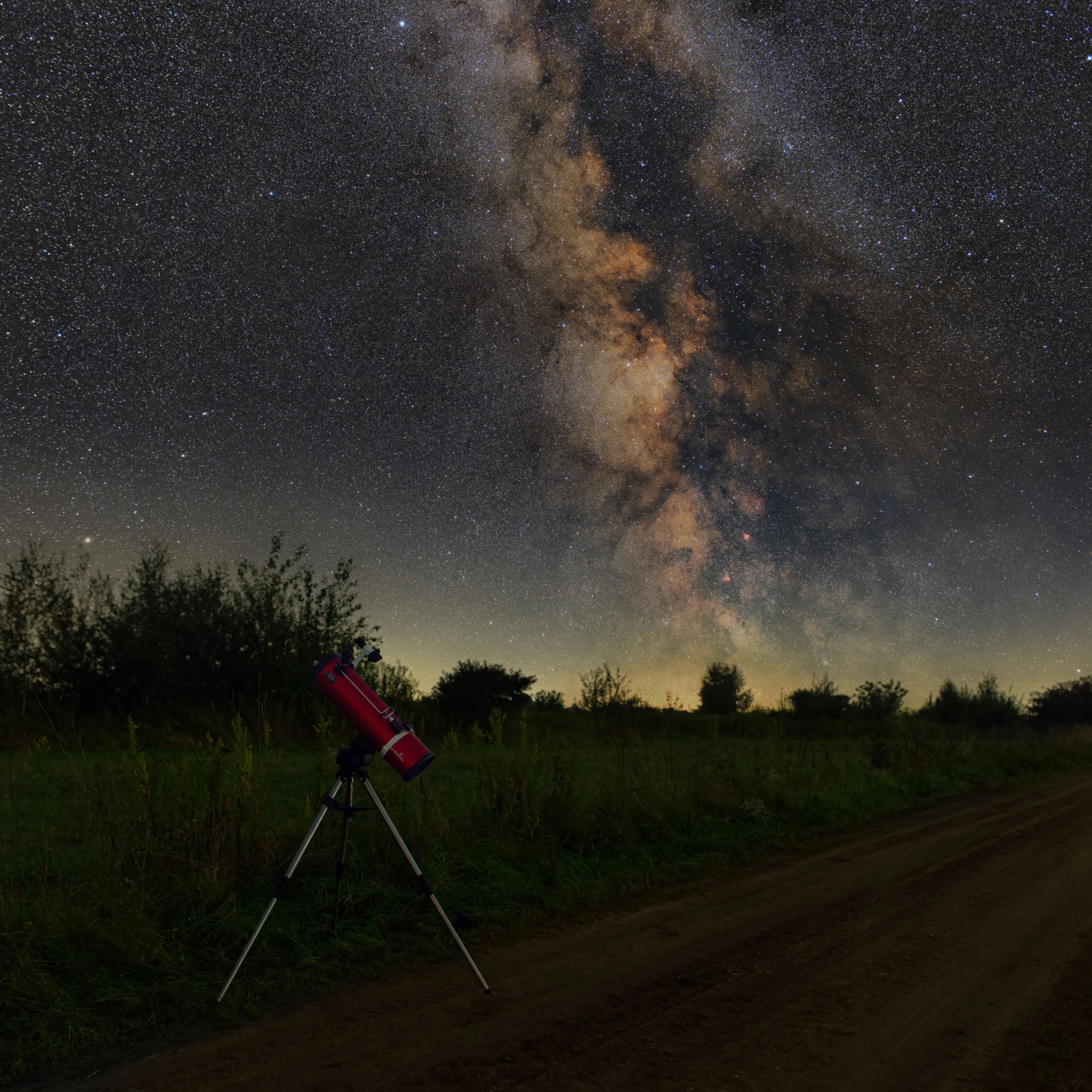 Silhouette of a red telescope on a tripod in a dark field with the Milky Way visible above the treeline.