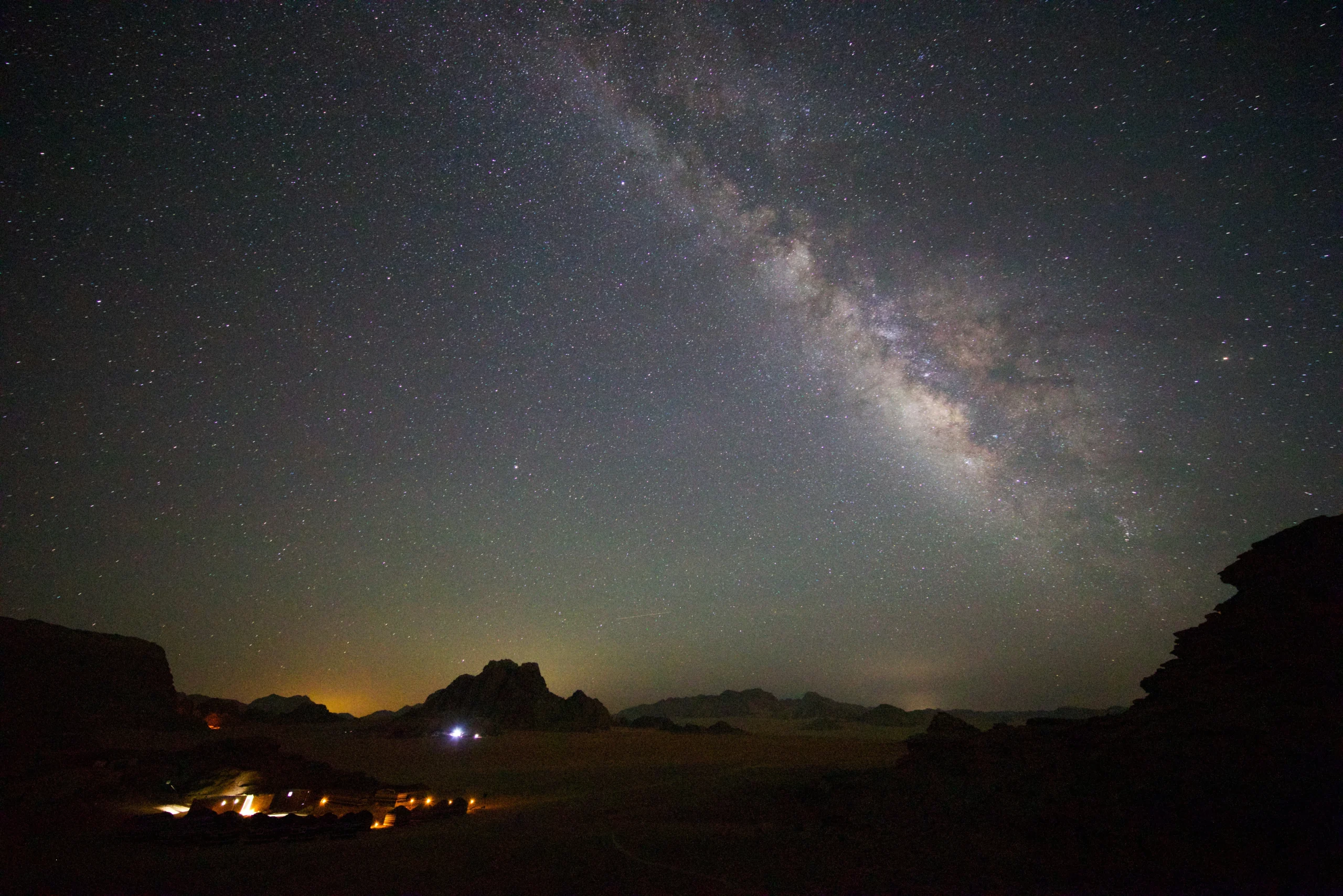 A dark desert landscape in Wadi Rum featuring a small, illuminated Bedouin camp at the base of rocky mountains, with the Milky Way glowing brightly overhead.