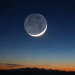 Thin crescent Moon with faint earthshine visible over a twilight mountain horizon.