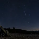Observer seated beneath a starry sky with Orion and a single meteor.