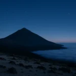 Volcanic cone rising above a dense layer of cloud at dusk with the first stars appearing.