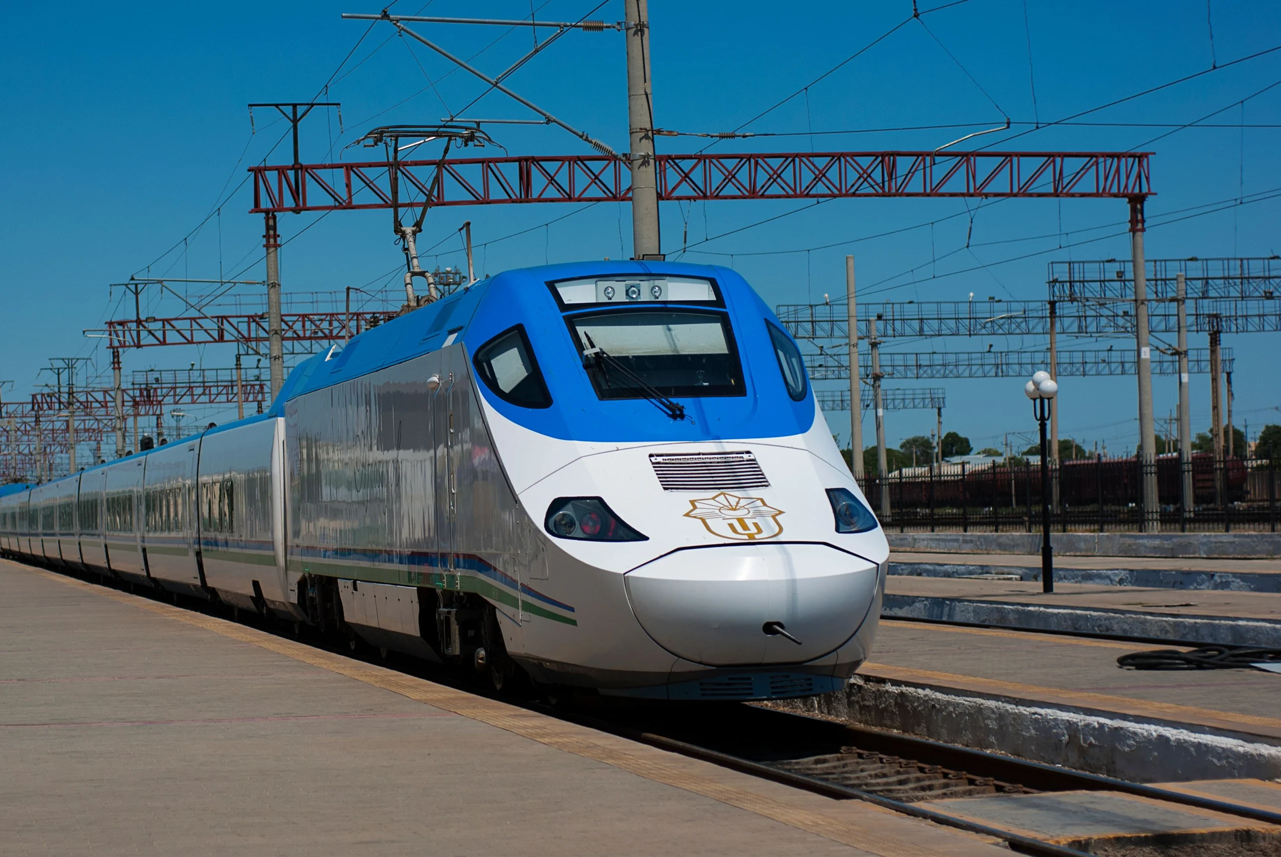 White high-speed passenger train on the tracks in Uzbekistan under a clear blue sky.