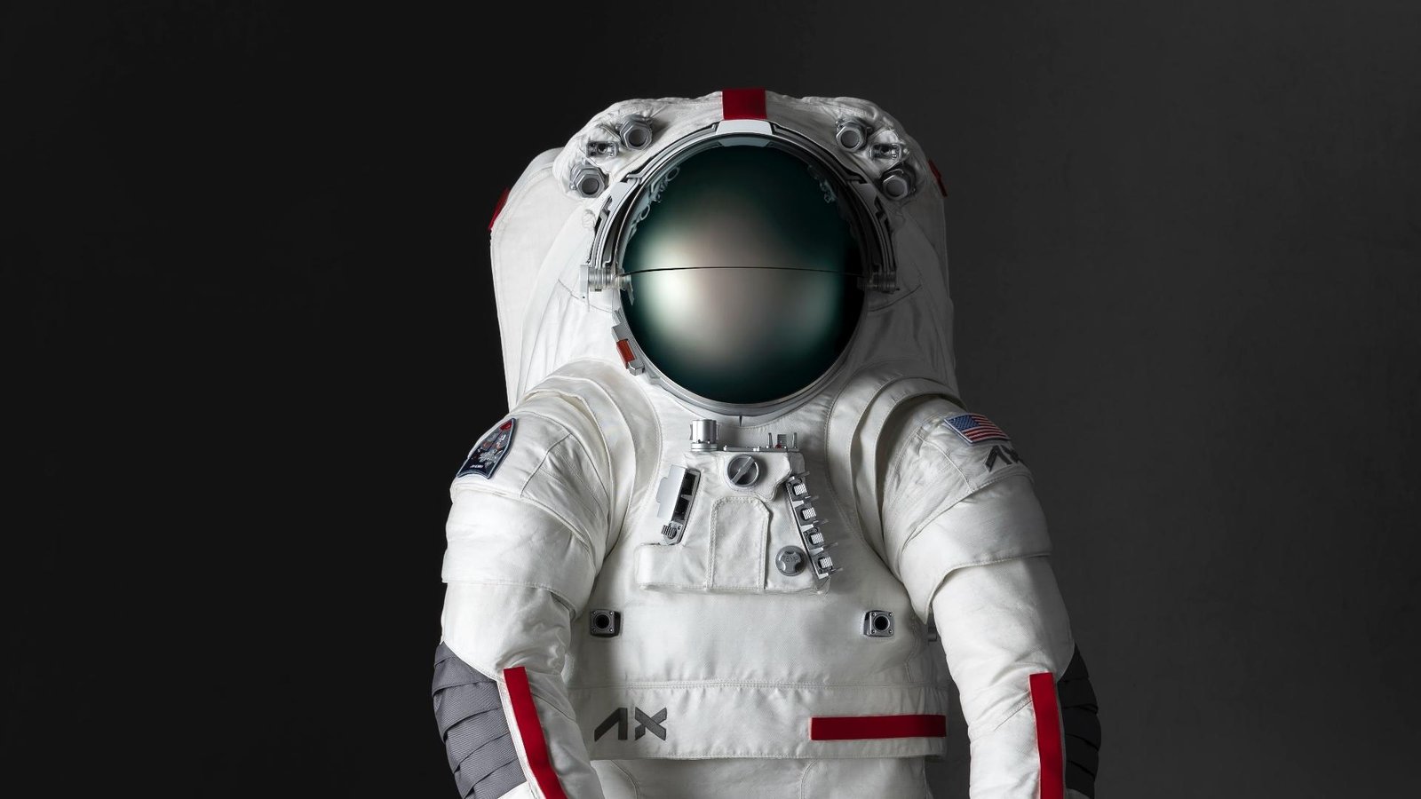A front-facing mockup of the white Axiom Space spacesuit designed for the Artemis 3 mission. The suit features a dark reflective visor, red accent stripes on the arms and helmet, an American flag patch on the left shoulder, and mechanical connectors on the chest.