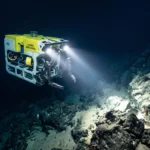 A yellow remotely operated vehicle illuminating the dark, rocky ocean floor of a deep-sea trench with twin headlights.