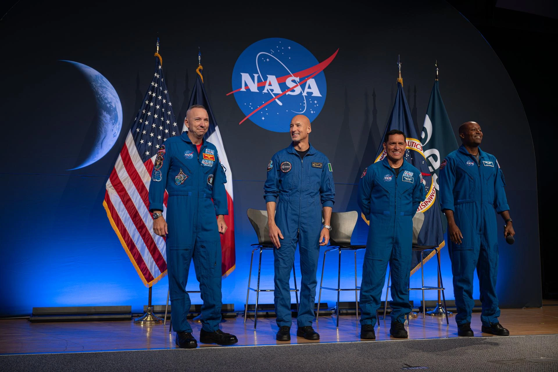 Four astronauts wearing blue flight suits smiling on a stage with a NASA backdrop and American flags during the Artemis III crew announcement.